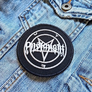 May include: A circular black patch featuring white embroidered text and a pentagram design. The patch is set against a denim jacket. The text is in a gothic font, possibly a band or brand name.