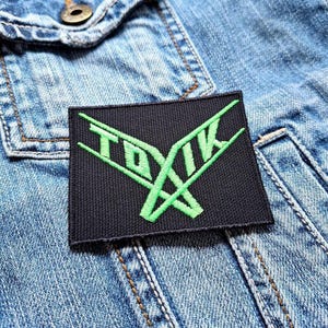 Peut inclure: Un écusson brodé noir avec le mot "TOXIK" en vert vif, avec un design stylisé. L'écusson est rectangulaire et est posé sur une veste en jean.