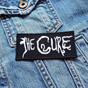 May include: A black rectangular patch with the white text "THE CURE" is sewn onto a denim jacket. The patch features stylized lettering, with the band's name prominently displayed. The denim jacket has a button and pocket detail.