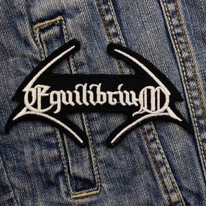 Könnte beinhalten: Schwarz-weiß gestickter Patch mit dem Wort "Equilibrium" in gotischer Schrift. Der Patch hat die Form eines Flügelpaares.