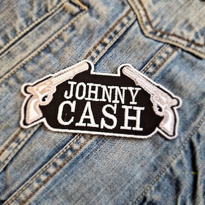 Pode incluir: Patch bordado preto e branco com o texto "JOHNNY CASH" e duas pistolas brancas de cada lado.