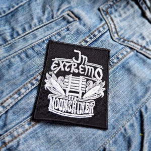 Può includere: Una toppa rettangolare nera con testo e immagini ricamate in bianco. Il testo recita "Jr. Extremo Moonshiner" con un disegno di mais e una struttura in legno. La toppa è posta su uno sfondo di denim blu.