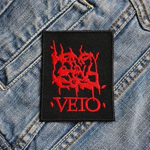 Könnte beinhalten: Schwarzer Patch mit roter Stickerei, die "Veto" lautet. Die Schrift ist in einer stilisierten Schriftart gehalten und von einem roten, tropfenden Design umgeben.