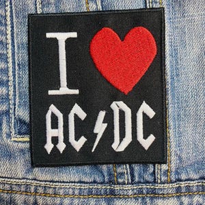 Op de afbeelding: Zwart vierkantige patch met de tekst "I ♥ AC/DC" in het wit en een rood hart. De patch is op een spijkerjas genaaid. De patch is een accessoire met een muzikaal thema.