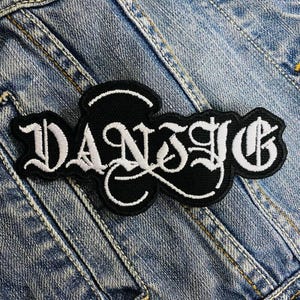 Op de afbeelding: Zwart en wit geborduurde patch met het woord "DANSIG" in een gotisch lettertype. De patch is rechthoekig en heeft een zwarte achtergrond met witte stiksels. De patch is op een spijkerjasje.