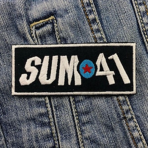 Könnte beinhalten: Schwarzer und weißer gestickter Patch mit dem Text "SUM 41" in Weiß mit einem blauen Kreis und einem roten Stern in der Mitte.