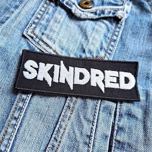 Könnte beinhalten: Schwarz-weiß gestickter Patch mit dem Wort "SKINDRED" in weißen Buchstaben auf schwarzem Hintergrund.