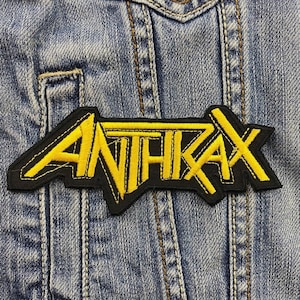 Może przedstawiać: Naszywka haftowana w kolorze czarnym i żółtym z napisem "ANTHRAX" w pogrubionym, stylizowanym kroju.