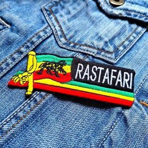 Può includere: Una toppa ricamata rossa, gialla e verde con un leone e le parole "Rastafari" su una giacca di jeans.