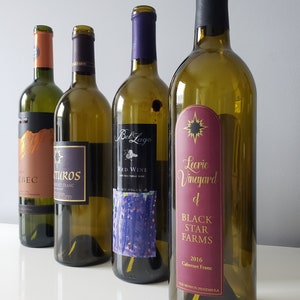 May include: Four empty wine bottles are displayed, each with a unique label. The labels feature text such as "Bel Lago Red Wine" and "Leorie Vineyard of Black Star Farms." The bottles are green glass, and the labels vary in color and design.