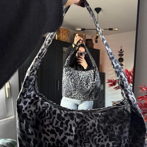 Könnte beinhalten: Eine schwarze und graue Schultertasche mit Leopardenmuster und Reißverschluss. Die Tasche wird von einer Person gehalten, die einen schwarzen Pullover trägt. Der Träger der Tasche ist ebenfalls mit schwarzen und grauen Flecken gemustert.