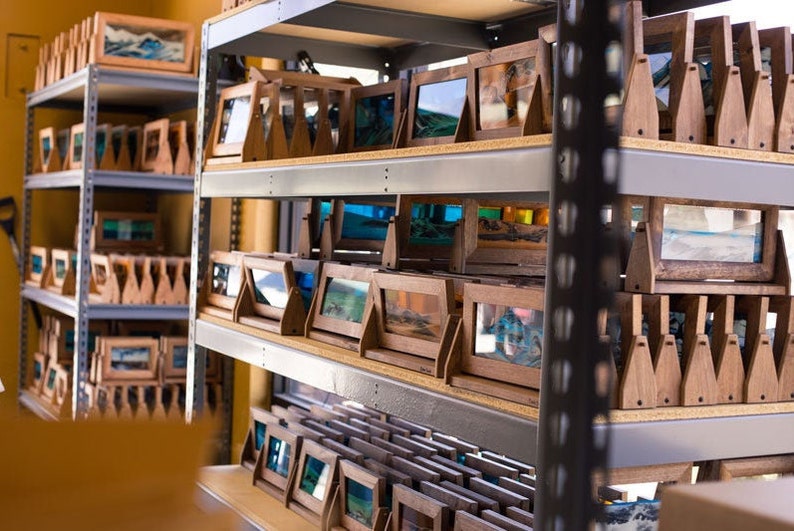 May include: A shelf unit filled with wooden frames containing sand art. The frames are arranged in rows and are all the same size. The sand art is a variety of colors and designs.