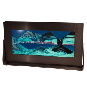 Op de afbeelding: Een zwart metalen frame omlijst een zandkunstwerk met blauw, groen en zwart zand. Het zand stroomt en creëert een uniek patroon.
