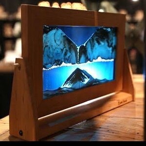 May include: A wooden frame holds a liquid sand art piece with blue sand and a clear liquid. The sand is flowing down from the top of the frame to the bottom. The frame has a light wood finish and a small, black switch on the side.