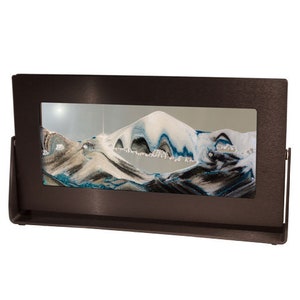 May include: A brown metal frame with a glass panel displaying a sand art design. The sand art features a white mountain range with blue and gray accents.