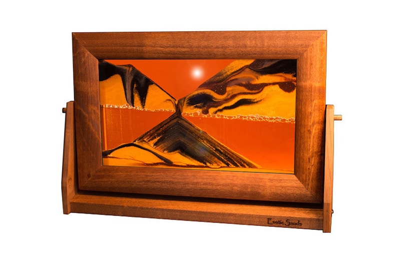 May include: A wooden framed sand art piece with a stand. The sand is orange and black and is flowing through the hourglass. The frame is dark brown and has the words "Exotic Sands" engraved on the bottom.