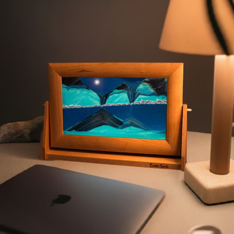 May include: A wooden frame sand art piece with a blue and green sand design. The frame is on a table with a laptop and a lamp.