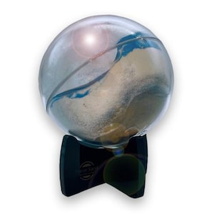 Könnte beinhalten: Eine klare Glaskugel gefüllt mit weißem und blauem Sand auf einem schwarzen Ständer. Der Sand wirbelt und erzeugt ein wellenförmiges Muster. Der Ständer trägt den Text "Planet Sand".