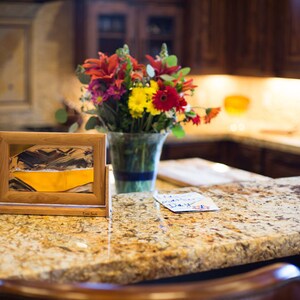 May include: A wooden frame with a sand art piece inside. The sand art is a yellow and brown landscape. The frame is sitting on a granite countertop with a bouquet of red, yellow, and orange flowers in a glass vase behind it.