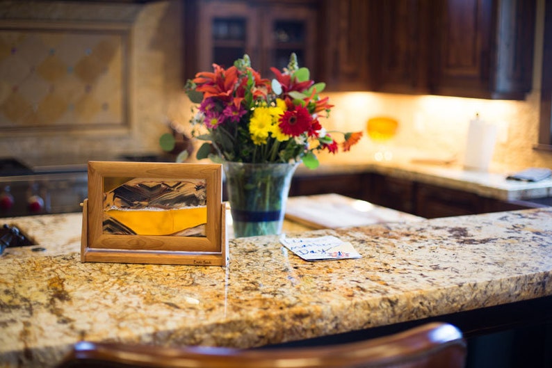 May include: A wooden frame with a sand art piece inside. The sand art is a yellow and brown landscape. The frame is sitting on a granite countertop with a bouquet of red, yellow, and orange flowers in a glass vase behind it.