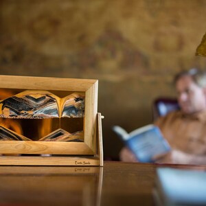May include: A wooden frame holds a sand art piece with a swirling pattern of black, brown, and gold sand. The sand art is on a table with a book and a lamp in the background.