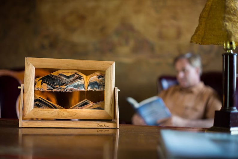 May include: A wooden frame holds a sand art piece with a swirling pattern of black, brown, and gold sand. The sand art is on a table with a book and a lamp in the background.