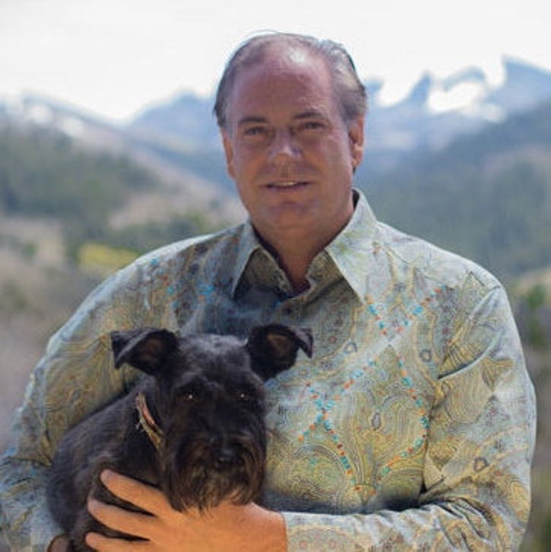 May include: A man in a light blue patterned shirt is holding a black schnauzer dog in his arms. The man is smiling and the dog is looking at the camera. The background is a blurry image of mountains.