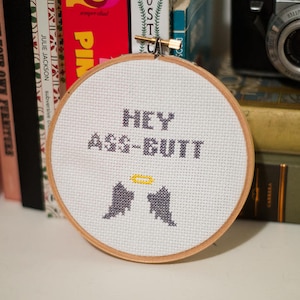 Könnte beinhalten: Ein Kreuzstich-Stickrahmen mit dem Text "HEY ASS-BUTT" in grauer Farbe. Darunter befinden sich zwei graue Engelsflügel und ein gelber Heiligenschein. Die Stickerei befindet sich in einem Holzrahmen.