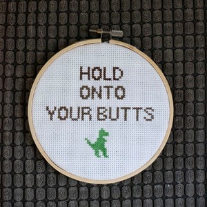 Puede incluir: Un bastidor de punto de cruz con la frase "HOLD ONTO YOUR BUTTS" en hilo marrón. Un dinosaurio verde está bordado debajo del texto. El bastidor está sobre un fondo gris texturizado.