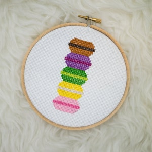 May include: A cross-stitch embroidery piece featuring a vertical stack of five colorful macarons in brown, purple, green, yellow, and pink, centered on a white fabric canvas held within a circular wooden embroidery hoop with a metal tightening screw.