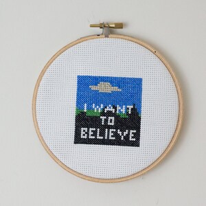 Könnte beinhalten: Kreuzstich-Stickerei in einem Holzreifen. Das Design zeigt einen blauen Himmel mit einer grauen UFO, grünen Boden und den Text "I WANT TO BELIEVE" in Schwarz und Weiß.