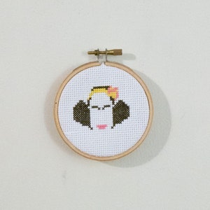 May include: A cross-stitch embroidery of a stylized face in a wooden hoop. The face features black hair, a yellow and pink headband, and a pink mouth. The embroidery is set against a white background.