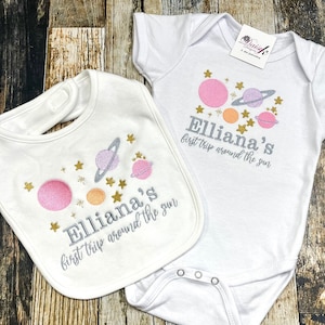 May include: A white baby bib and onesie with a space theme. The bib and onesie feature a silver glitter design of planets, stars, and the text "Elliana's first trip around the sun".
