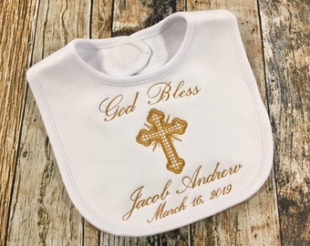 May include: White bib with gold embroidery. The embroidery says "God Bless Jacob Andrew March 16, 2019" and features a cross.