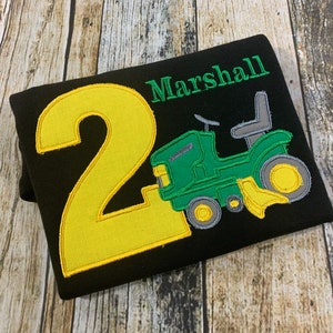 May include: Black t-shirt with a yellow number 2 and a green tractor with a gray seat. The text "Marshall" is embroidered in green above the tractor.