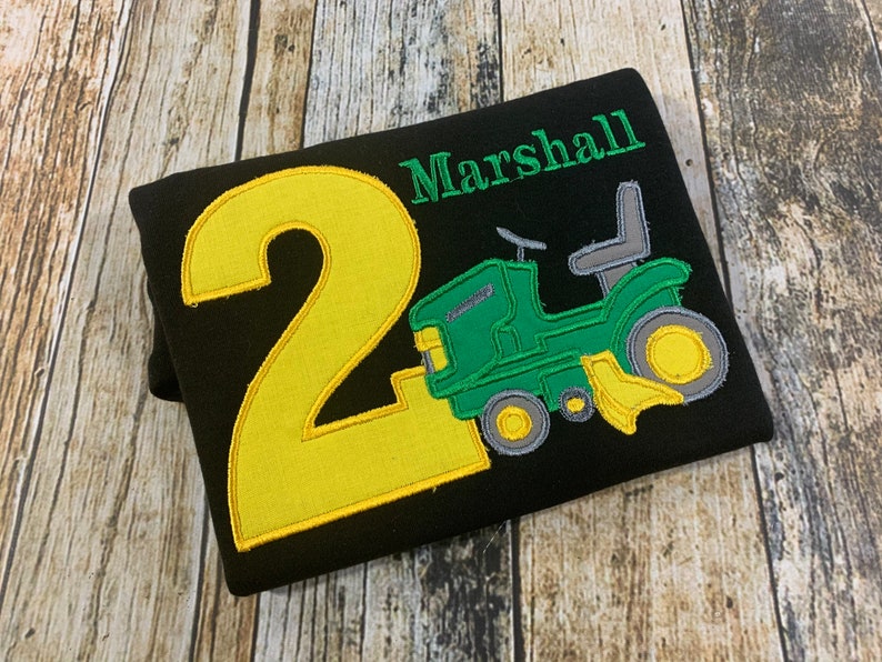 May include: Black t-shirt with a yellow number 2 and a green tractor with a gray seat. The text "Marshall" is embroidered in green above the tractor.