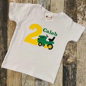 May include: White t-shirt with a yellow number 2 and a green tractor. The text "Caleb" is embroidered on the shirt.