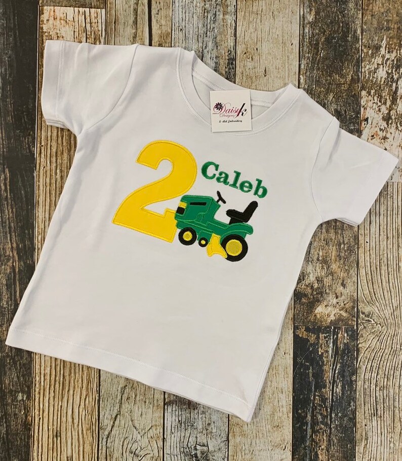 May include: White t-shirt with a yellow number 2 and a green tractor. The text "Caleb" is embroidered on the shirt.