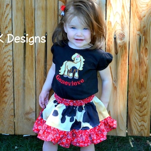 May include: A young girl wearing a black t-shirt with a brown horse and the name "Genevieve" embroidered on it. She is also wearing a multi-colored skirt with a cow print pattern and a red and white ruffle trim.
