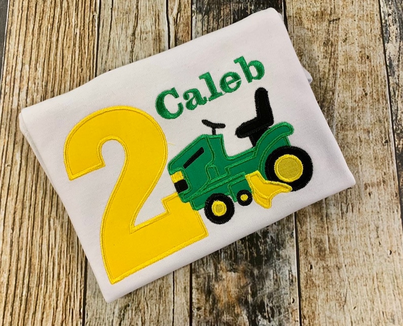 May include: A white t-shirt with a yellow number 2 and a green tractor with a black seat. The name Caleb is embroidered in green above the tractor.