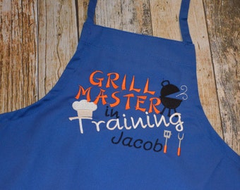 May include: Blue apron with the text "Grill Master in Training Jacob" embroidered in white, orange, and black. The design includes a chef's hat, a grill, a spatula, and a fork.