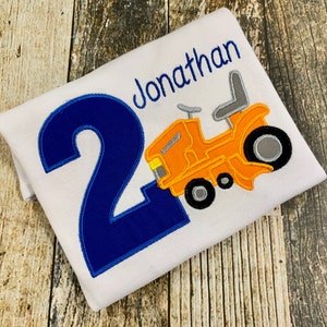 May include: A white t-shirt with a blue number "2" and a yellow tractor with a gray seat. The shirt also has the name "Jonathan" embroidered on it.