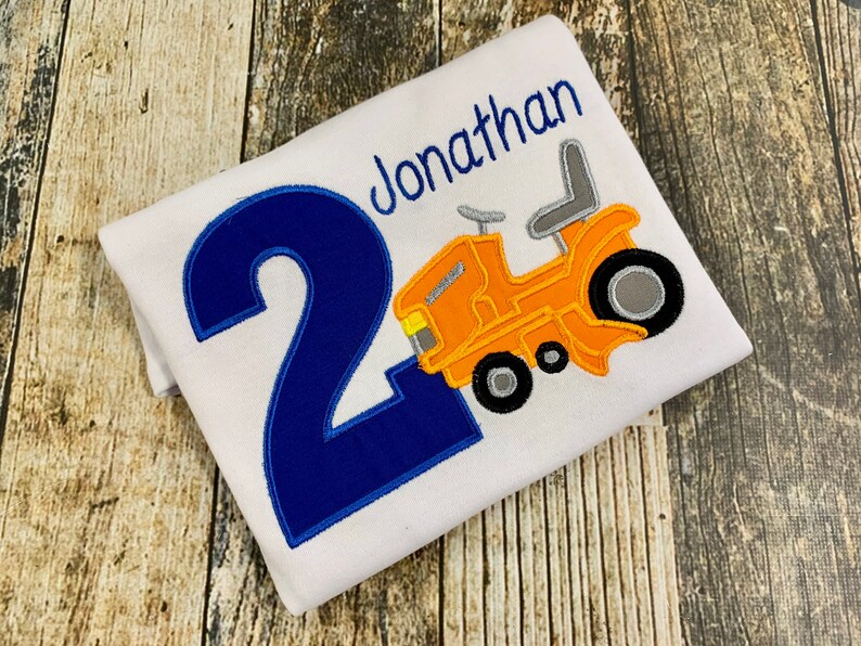 May include: A white t-shirt with a blue number "2" and a yellow tractor with a gray seat. The shirt also has the name "Jonathan" embroidered on it.