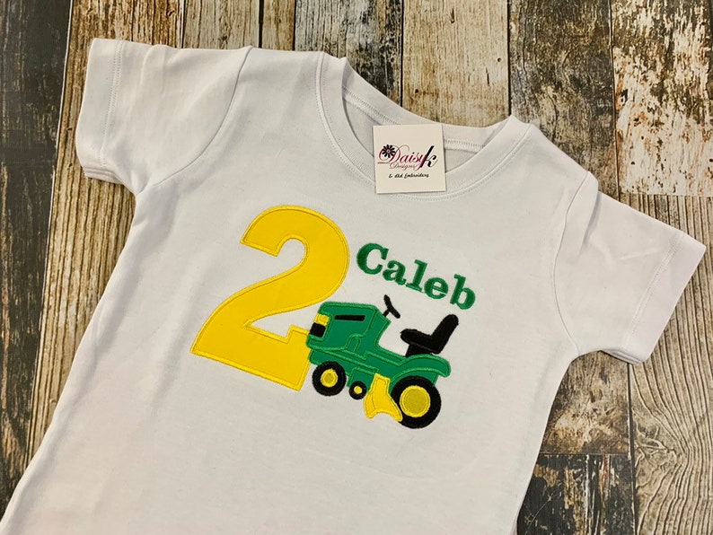 May include: White t-shirt with a yellow number 2 and a green tractor with the name Caleb embroidered on it.