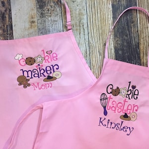 May include: Two pink aprons with embroidered cookie-themed designs. One apron says "Cookie Maker Mom" and the other says "Cookie Taster Kinsley".