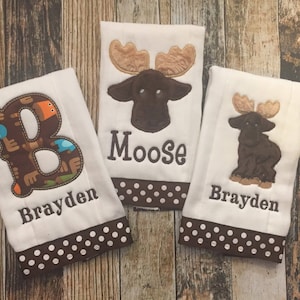 May include: Three white baby bibs with brown polka dot trim. One bib features a large brown letter "B" with animal designs, the second has a moose head, and the third has a moose silhouette. All bibs have the name "Brayden" embroidered.