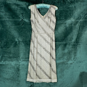 May include: A white sleeveless dress with a V-neckline and a silver beaded pattern. The dress is hanging on a hanger against a teal green background.