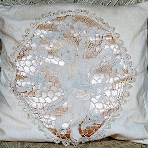 May include: A white pillow with a delicate lace design featuring a floral motif and a figure of an angel. The lace is a light beige color and the angel is a light blue color.