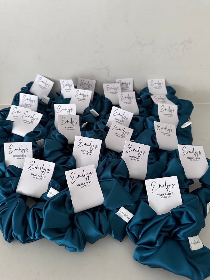 May include: Teal satin scrunchies with white labels that say "Emily's Hens Party 25-05-24".