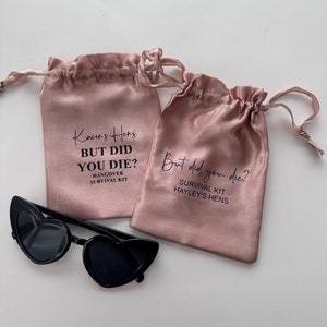 Peut inclure: Deux sacs en satin rose à cordon avec le texte "Kacie's Hens BUT DID YOU DIE? HANGOVER SURVIVAL KIT" et "But did you die? SURVIVAL KIT HAYLEY'S HENS" imprimé dessus.
